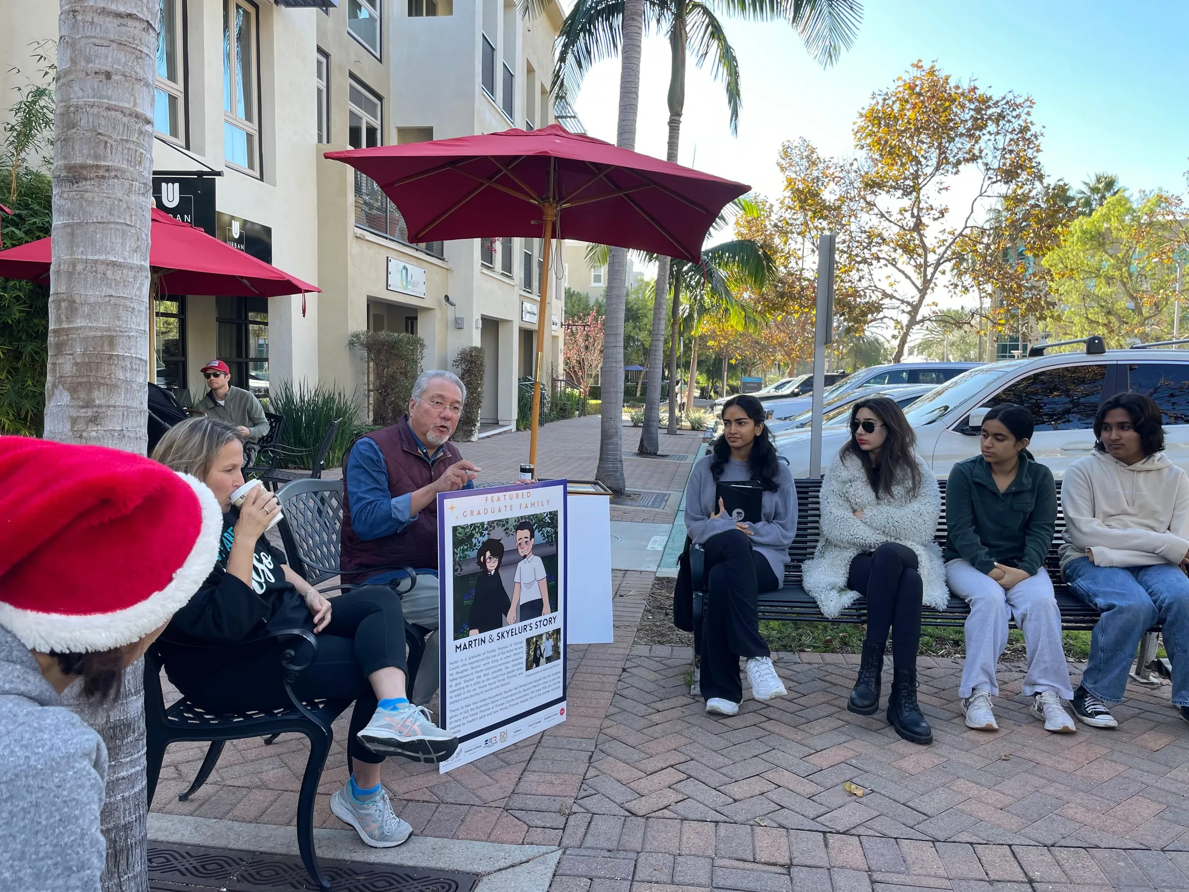 An outdoor meeting on a café patio where a man seated under a red umbrella presents a poster headed "Martin & Skyelur's Story" to members sitting on a bench opposite, one person nearby wearing a Santa hat