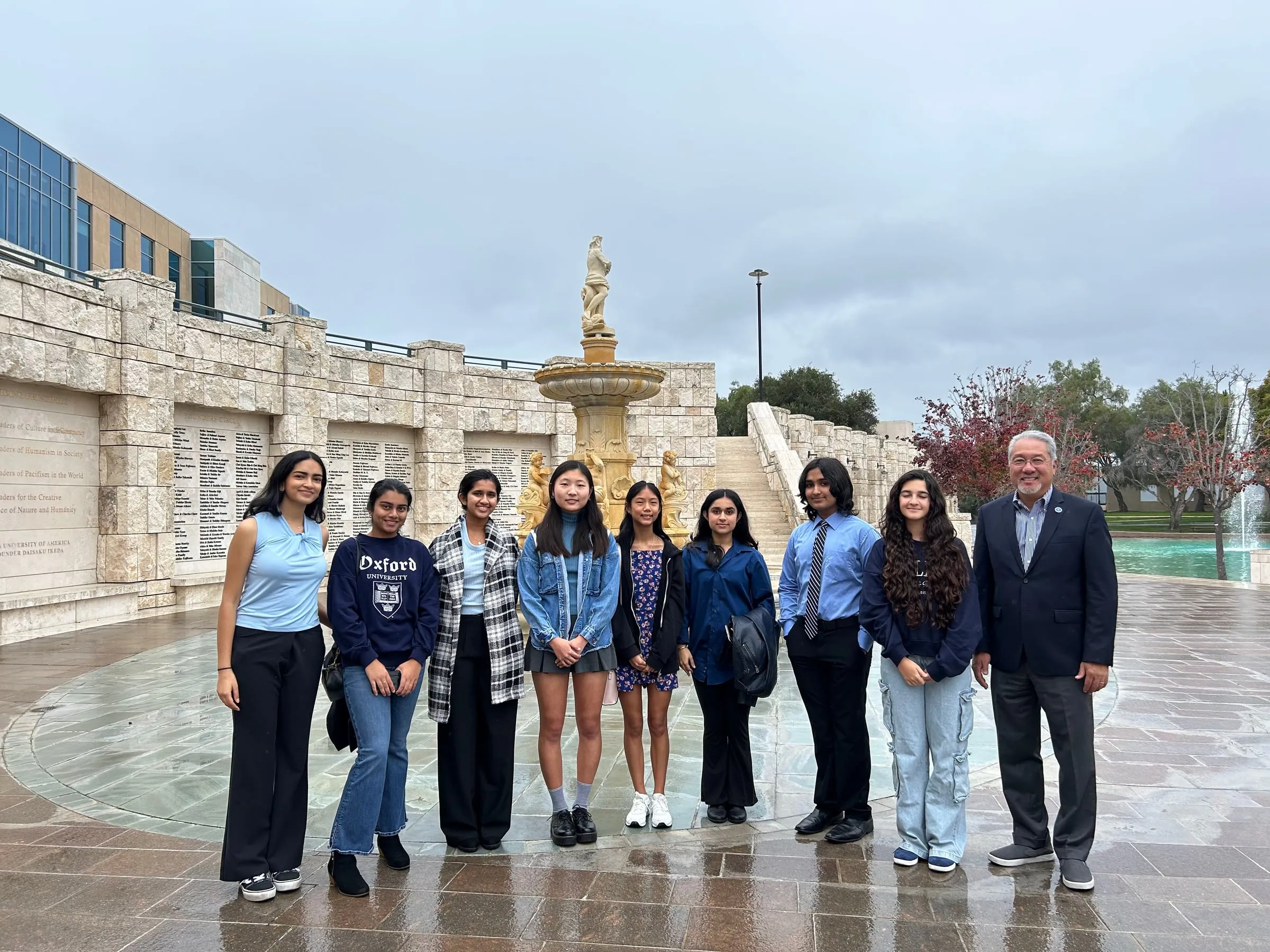 Nine council members and their advisor stand together on wet paving in front of the stone Founders Circle fountain at Soka University under a grey, overcast sky