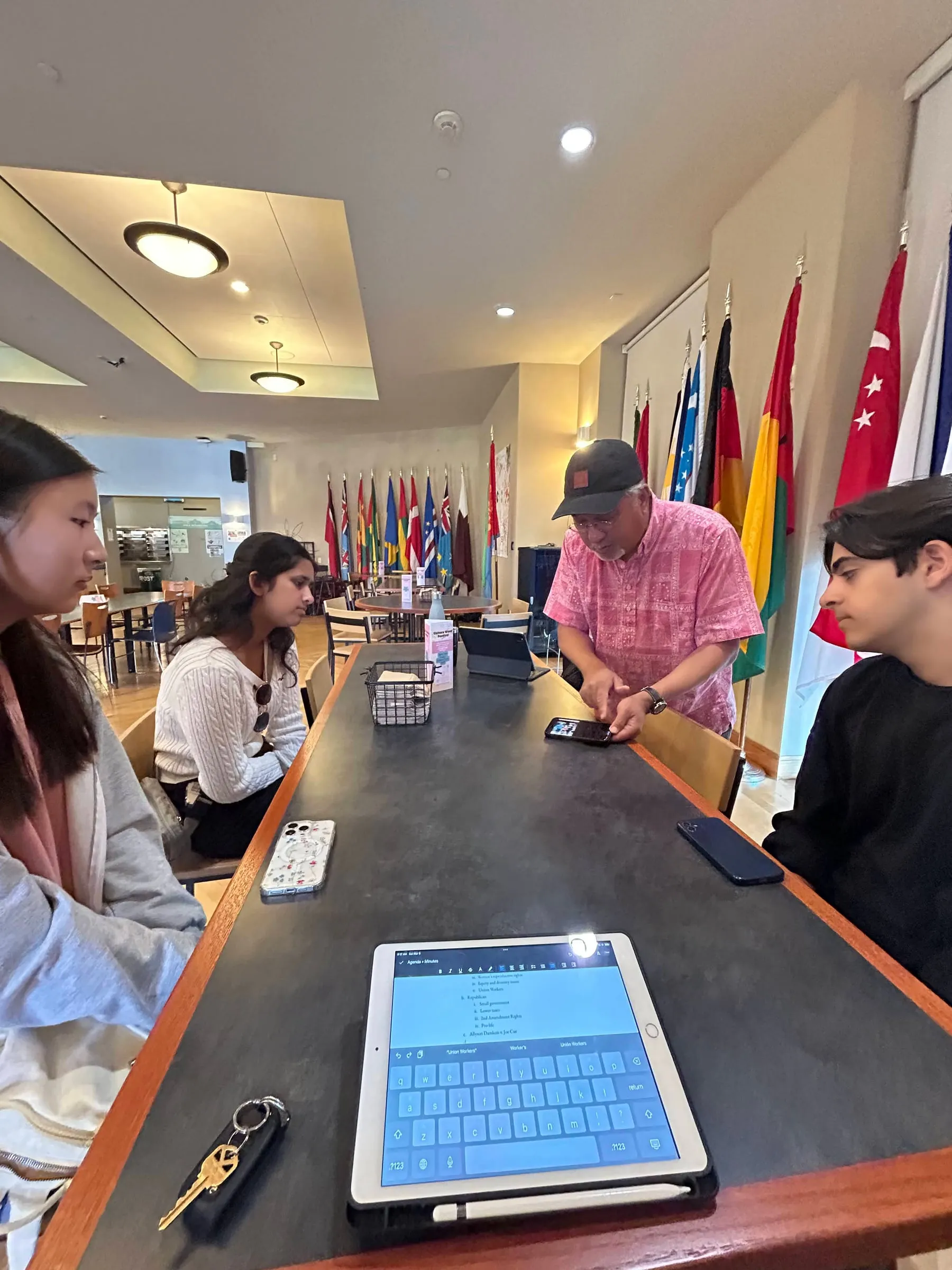 Members sit along a long counter in a dining hall hung with international flags while the council's advisor leans in to point at a phone, an iPad showing meeting minutes in the foreground