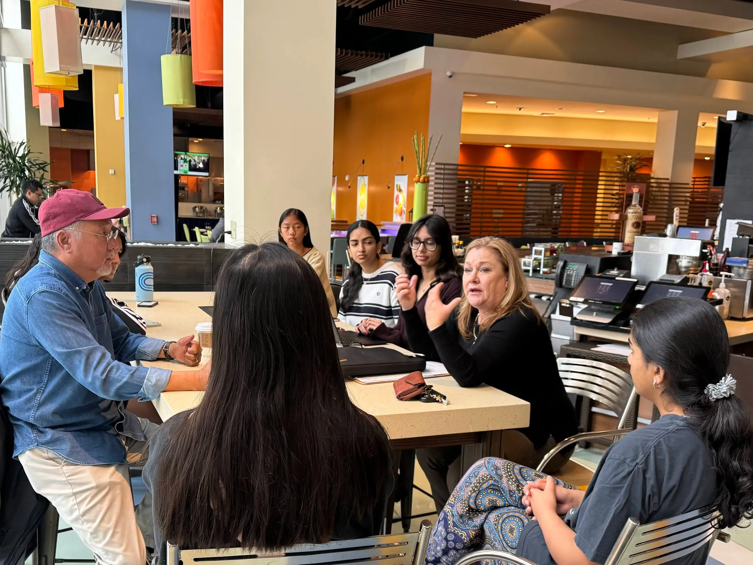 Council members gathered around a high table in a hotel restaurant listening to a guest speaker who is gesturing as she talks, with the council's advisor seated at the left