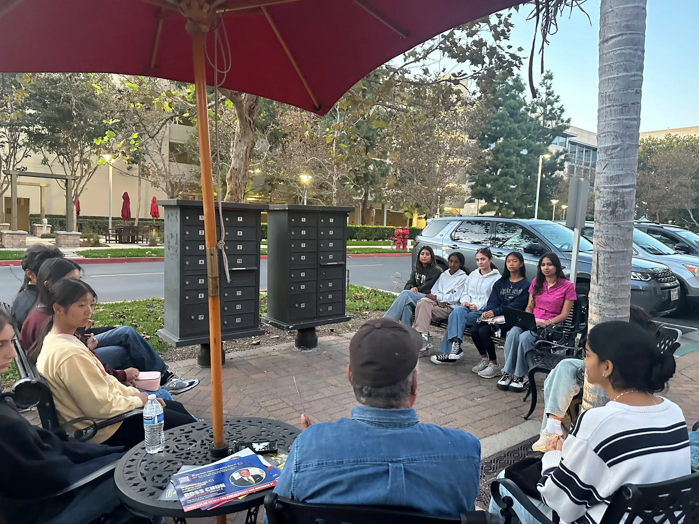 Council members sit in a circle of patio chairs under a red umbrella at an outdoor meeting, with a Ross Chun campaign mailer lying on the table in the foreground