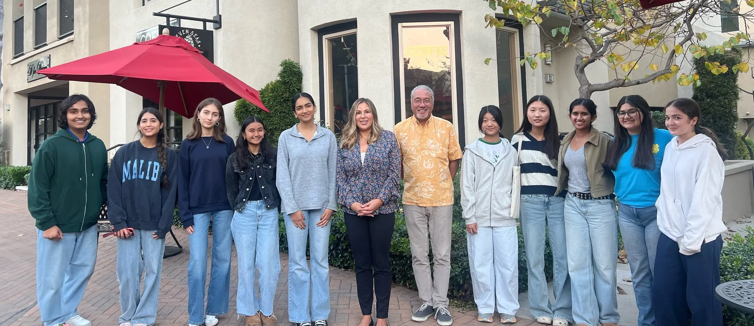 A wide group photo of council members standing on a brick patio outside a coffee shop, with the guest speaker and the council's advisor in the centre of the row