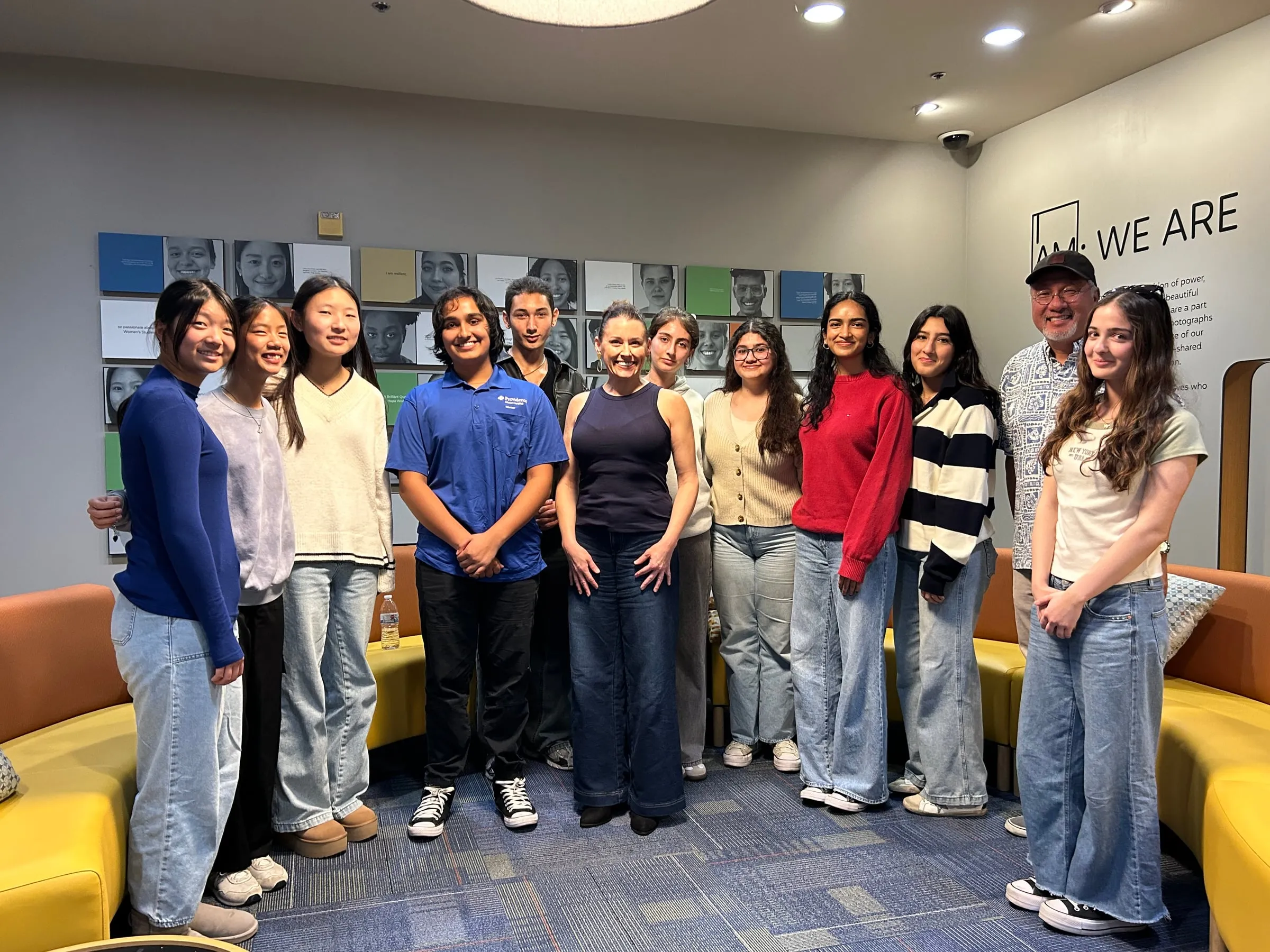 Twelve council members and their advisor line up for a group photo in a campus lounge in front of a wall of student portraits, with their guest speaker standing in the middle