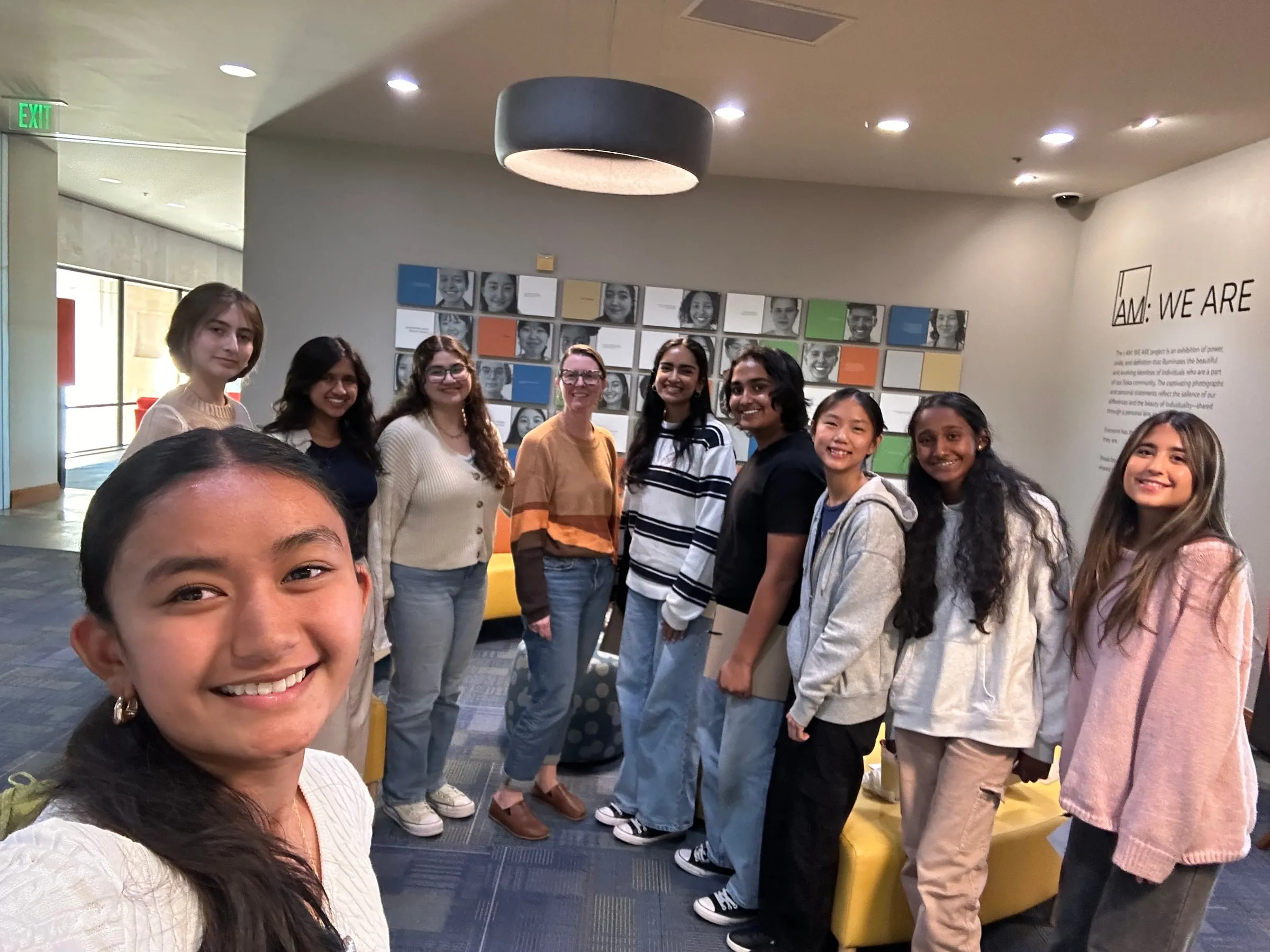 Council members crowd in for a group selfie in a Soka University lounge in front of the "I AM: WE ARE" portrait wall, with their guest speaker in an orange and brown striped sweater standing among them