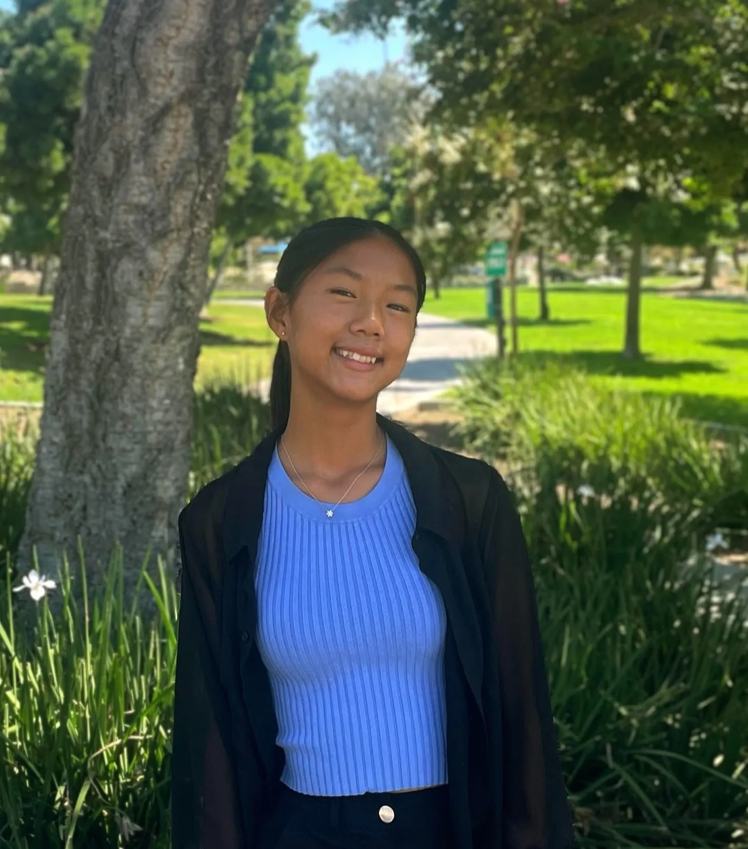 Catie Lacanienta in a park beside a tree, wearing a light blue ribbed top under an open black shirt
