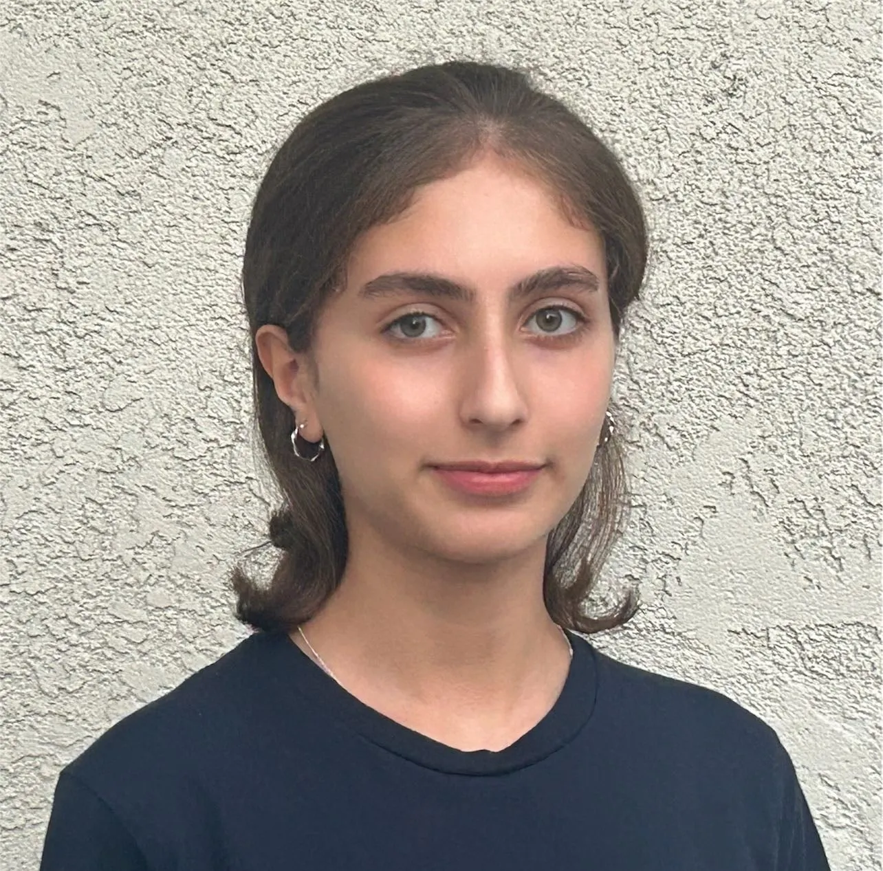 Diba Behbudi in a head-and-shoulders portrait against a textured cream stucco wall, wearing a navy crew-neck T-shirt