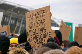 Protesters packed into an airport terminal, one holding a cardboard sign reading “No hate, no fear, Muslims are welcome here”