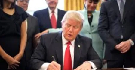 President Donald Trump signing a document at a desk in the Oval Office while officials stand behind him