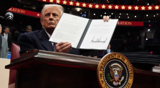 President Donald Trump holding open a signed executive order toward a crowd at an indoor inauguration-day event