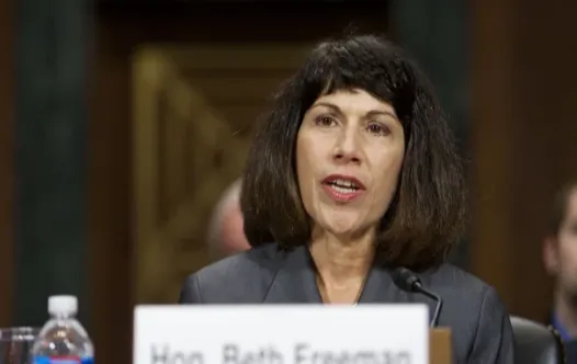 A judge speaking into a microphone at a hearing, with a nameplate in front of her reading “Hon. Beth Freeman”