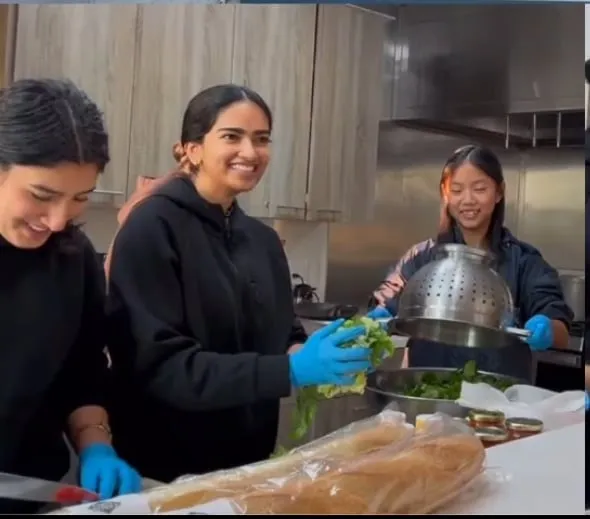 Three volunteers in disposable gloves prepare food in a stainless steel commercial kitchen, washing salad leaves into a colander beside loaves of bread