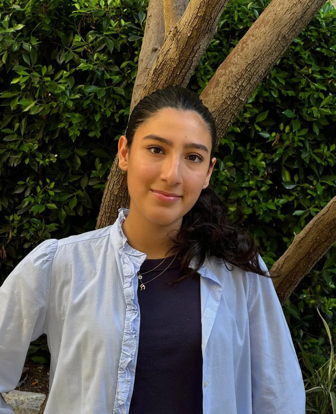 Hana Moradi beside a tree trunk in front of a dark hedge, wearing a pale blue ruffled shirt over a navy top