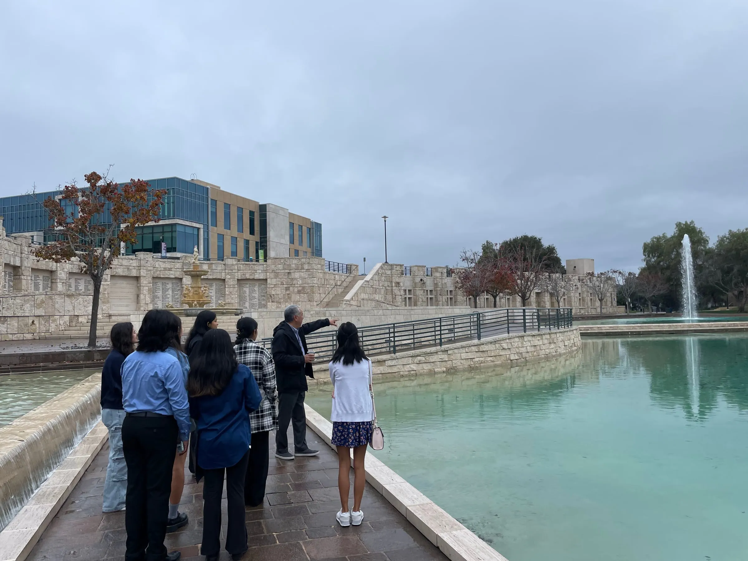 Council members stand at the edge of a long reflecting pool at Soka University while their advisor points across the water towards a stone fountain and the campus buildings beyond