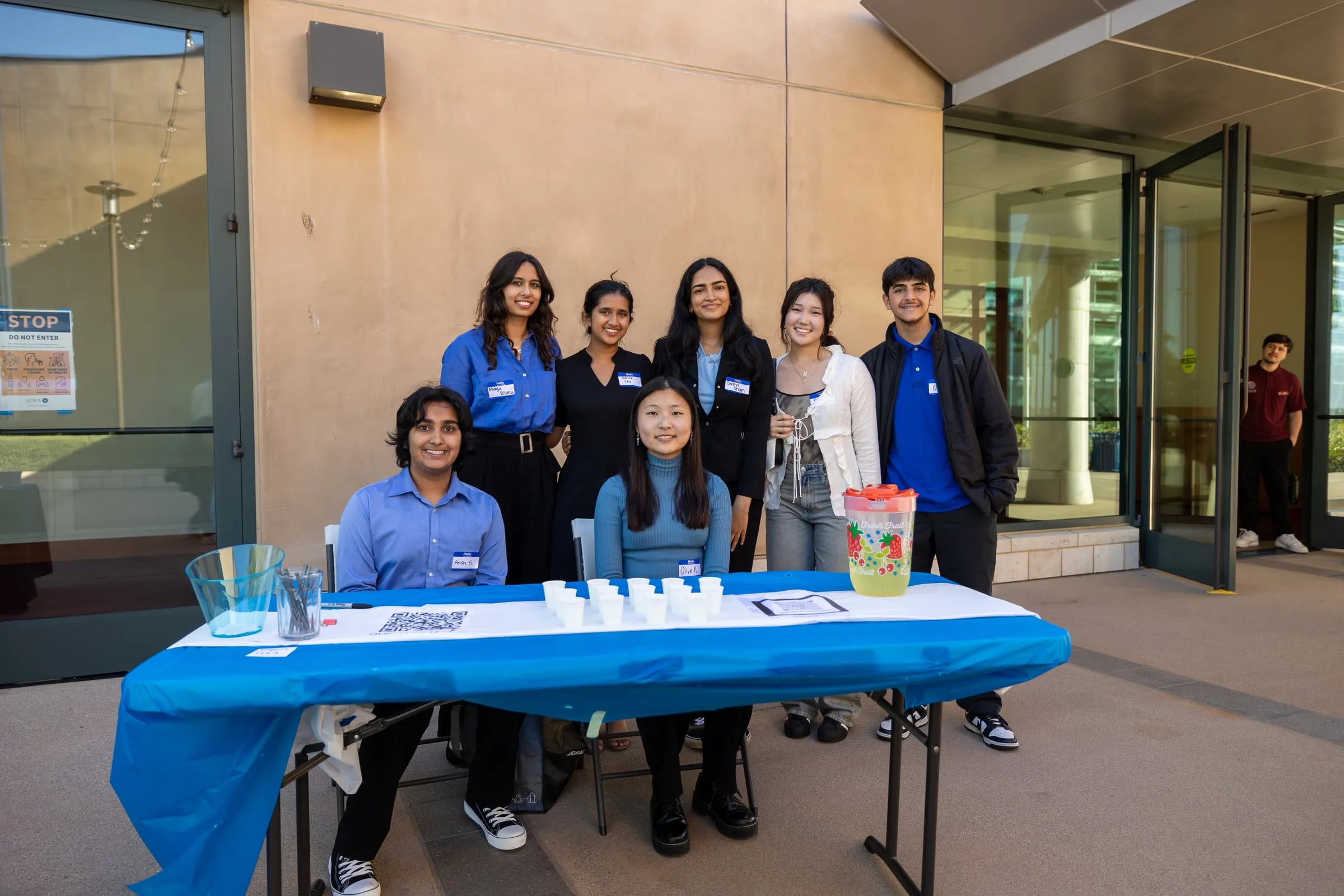 Seven council members wearing name badges pose at the outdoor check-in table, two seated behind a blue-draped table set with a QR code sign, cups and a drinks dispenser
