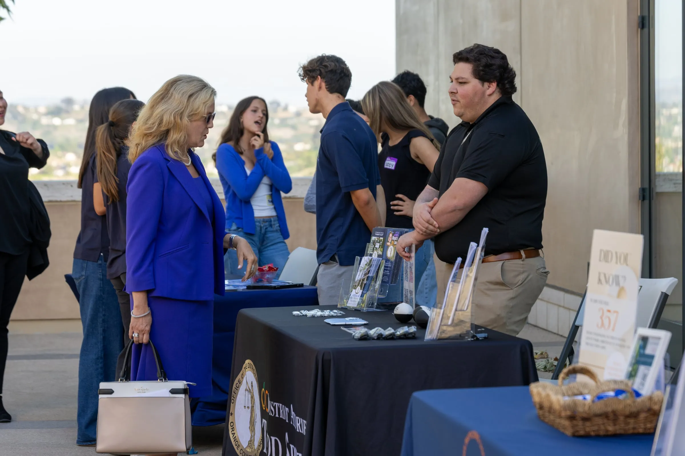 A woman in a bright blue suit stops at the District Attorney's outreach table on the terrace to talk with the volunteer staffing it, with attendees mingling behind