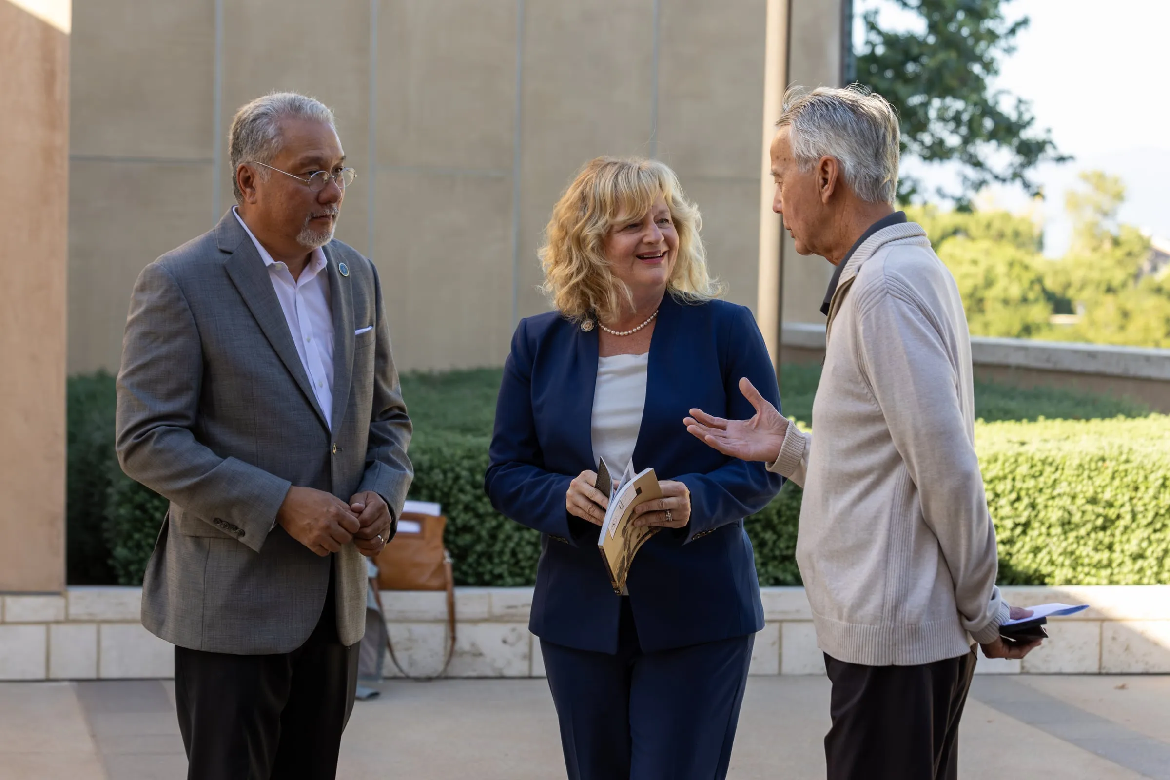 Three adults stand talking outside before the forum, one holding an open programme, in front of a low hedge and pale stone wall