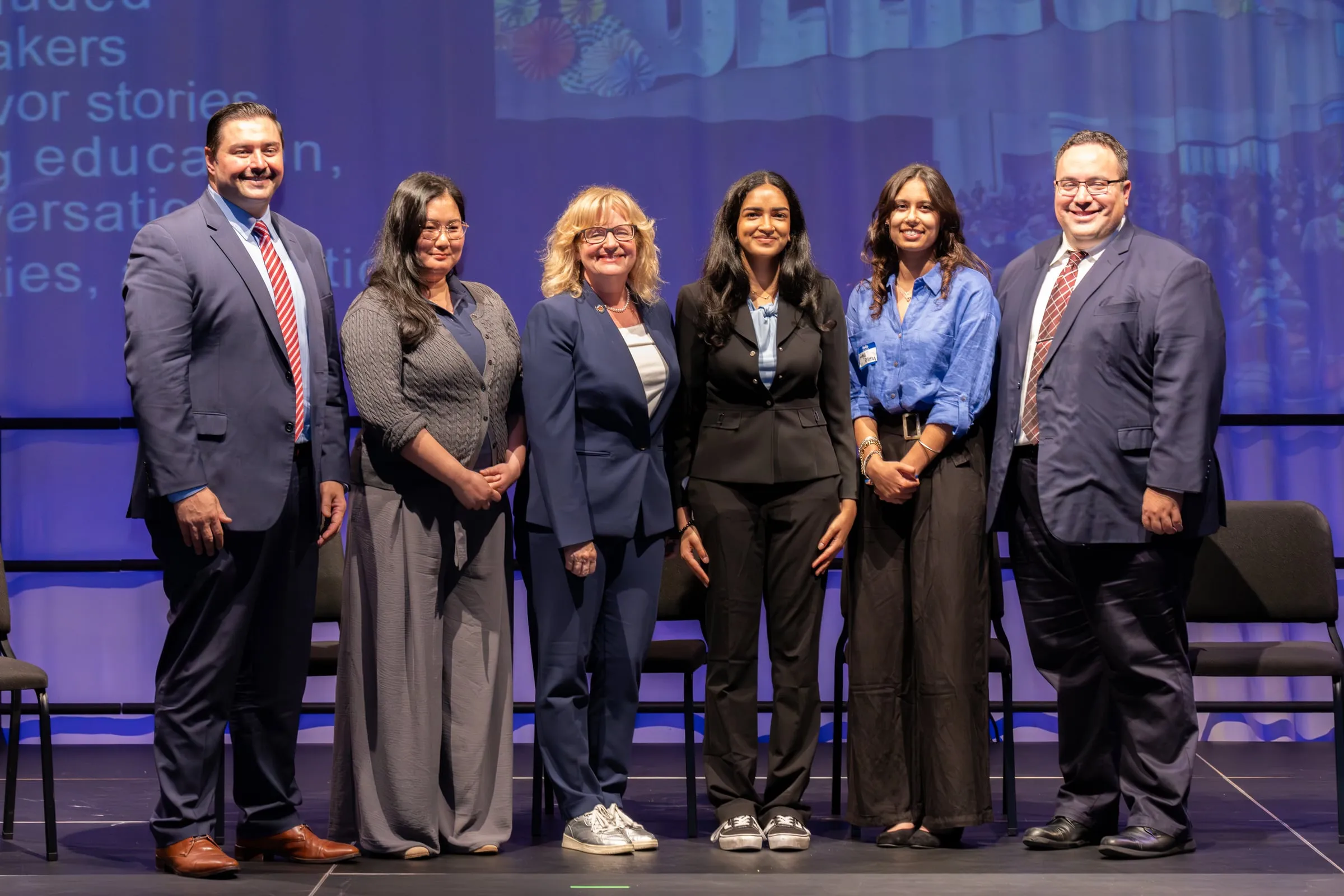 Six speakers and organisers stand in a row on stage for a photograph, lit blue by the projected slide behind them