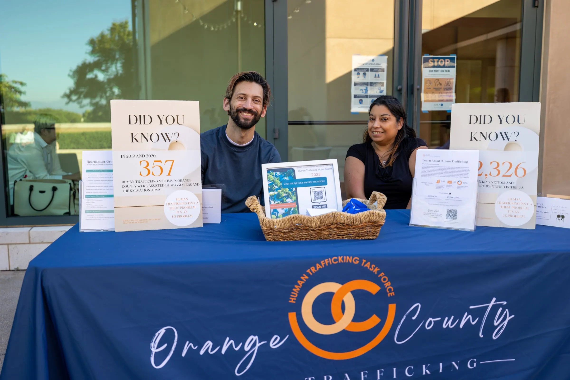 Two people sit behind the Orange County Human Trafficking Task Force resource table, flanked by "Did You Know?" display boards quoting victim statistics