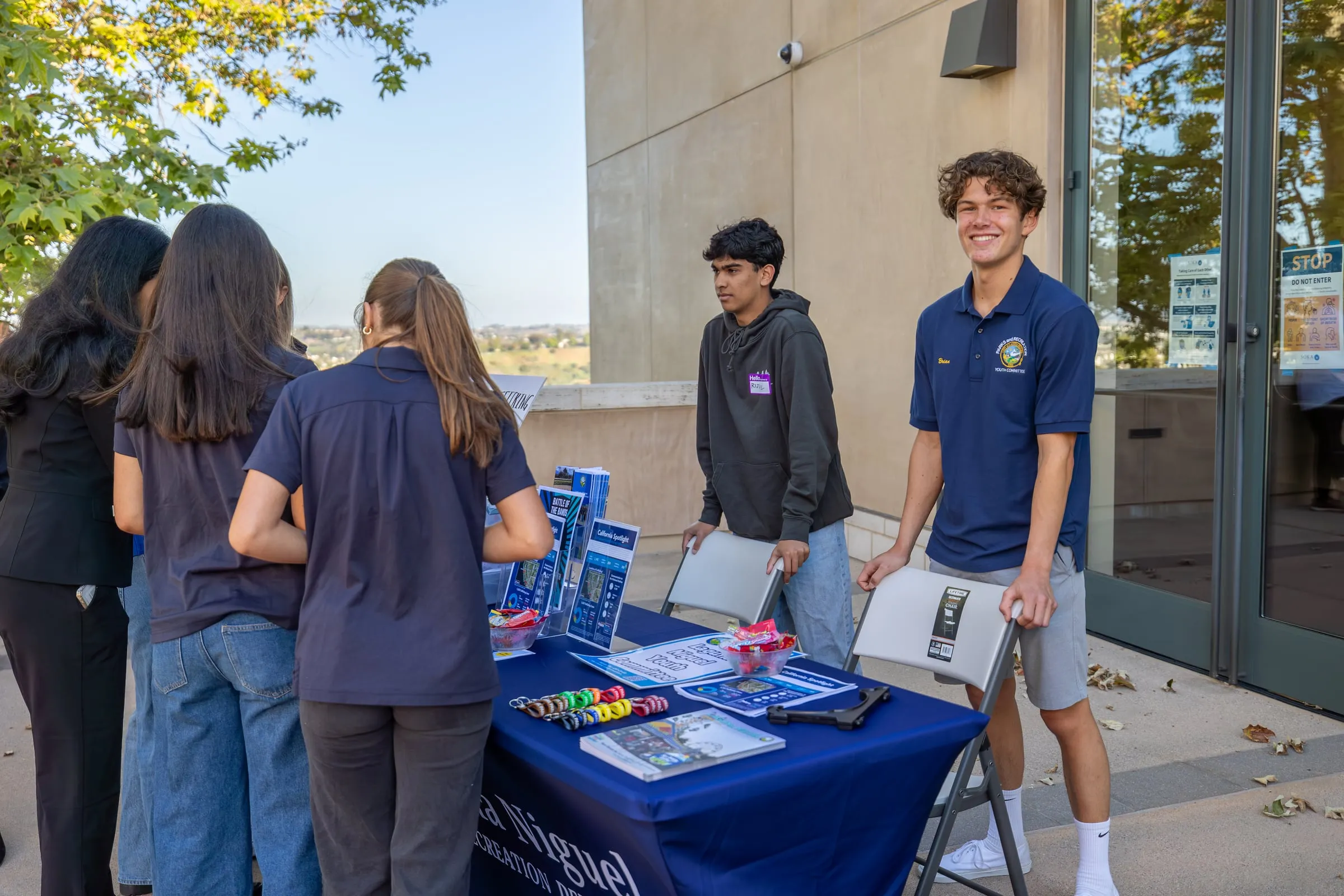 Teenagers in navy Parks and Recreation Youth Committee polo shirts set up the Laguna Niguel resource table outside, unfolding chairs and arranging leaflets and keychains