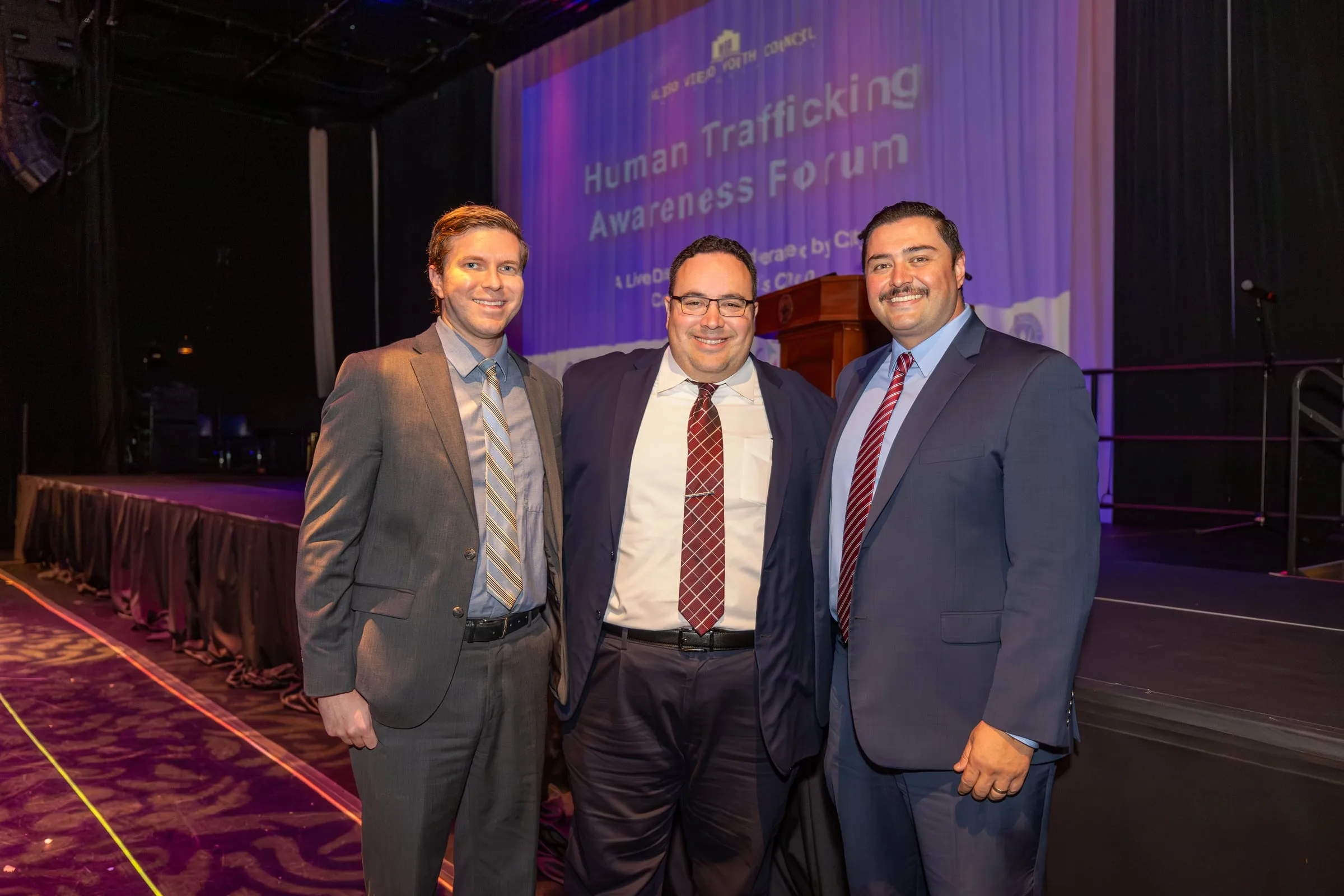 Three men in suits stand beside the stage in front of the projected title slide reading "Human Trafficking Awareness Forum"