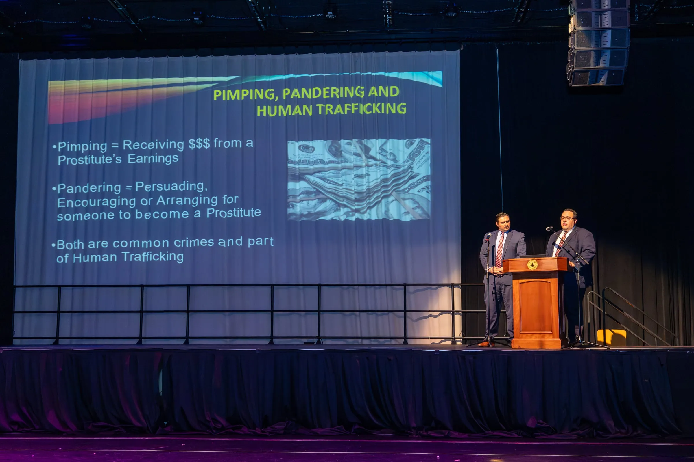 Two investigators speak from the lectern at the side of the stage while a slide headed "Pimping, Pandering and Human Trafficking" fills the screen beside them