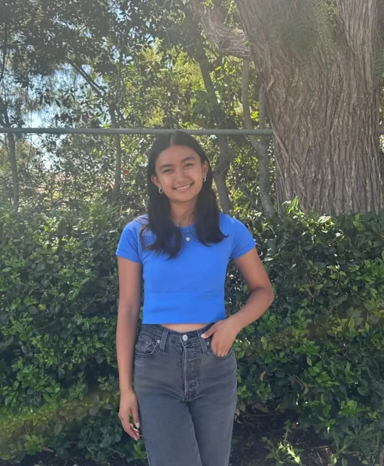 Jayzelle Jahanzeb standing in front of a hedge and a large tree, wearing a blue T-shirt and grey jeans with one hand in her pocket