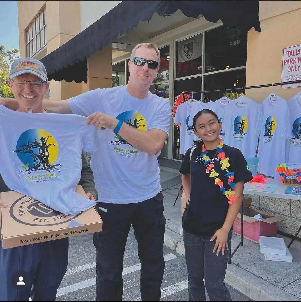 A council member wearing a flower lei stands outside a pizzeria beside two men holding up white "Music for Maui" T-shirts, with more shirts on hangers and a donation table behind them