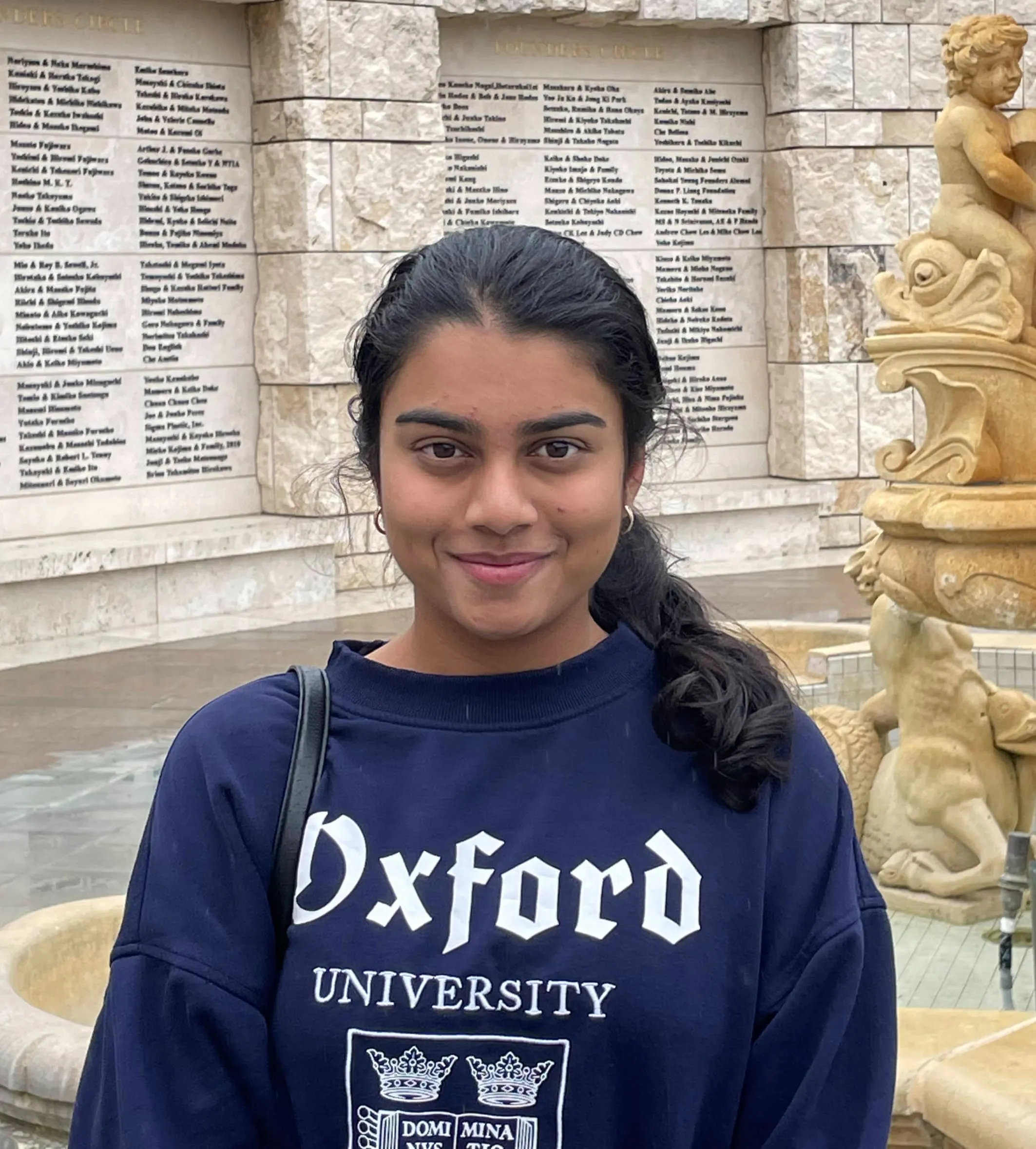 Anika Chandran in a navy Oxford University sweatshirt, standing by the Founders Circle donor wall and fountain