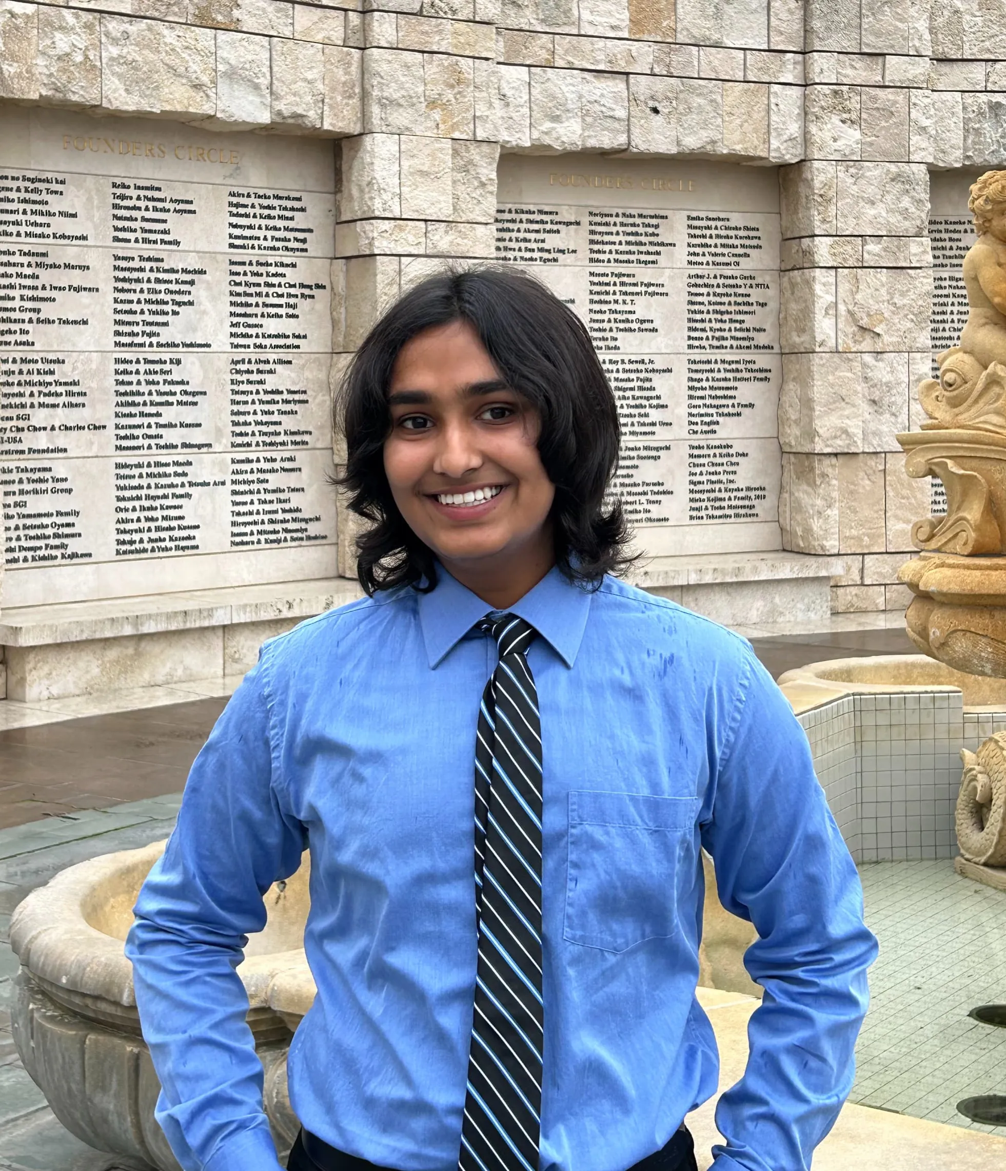 Ansh Sharma in a blue dress shirt and striped tie, standing in front of the Founders Circle donor wall