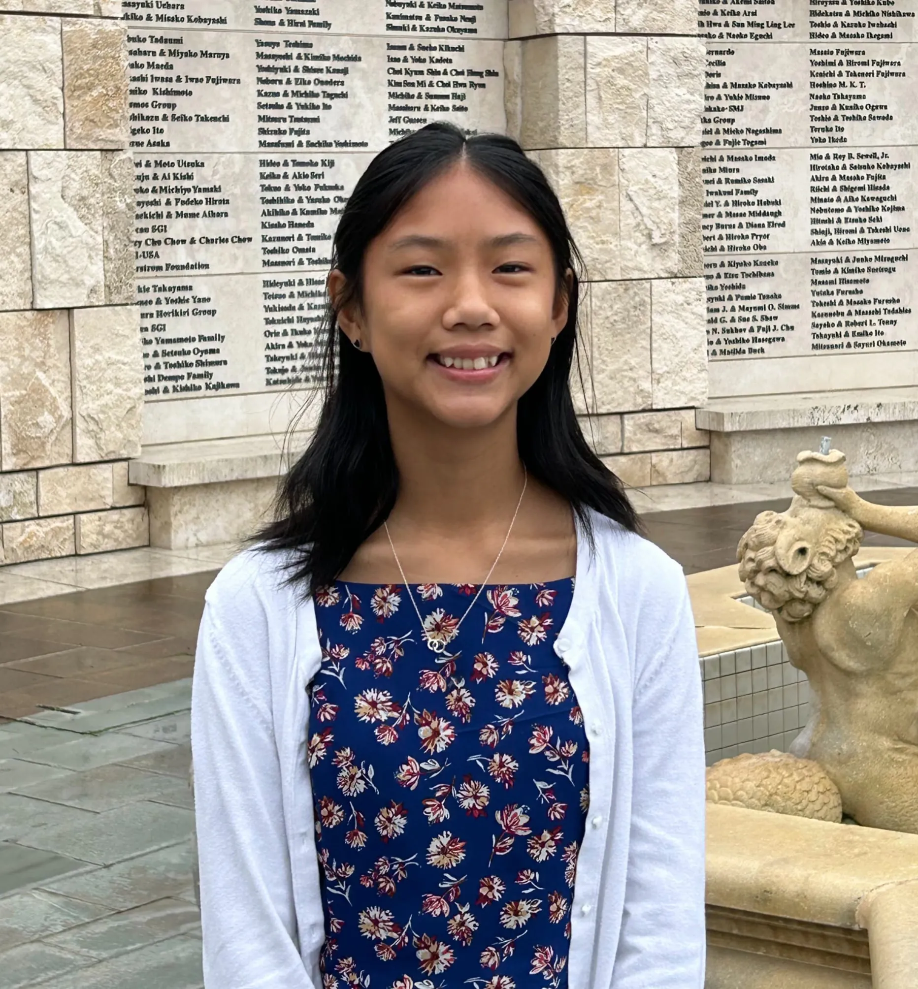 Catherine Lacanienta in a navy floral dress and white cardigan, standing in front of an inscribed stone donor wall