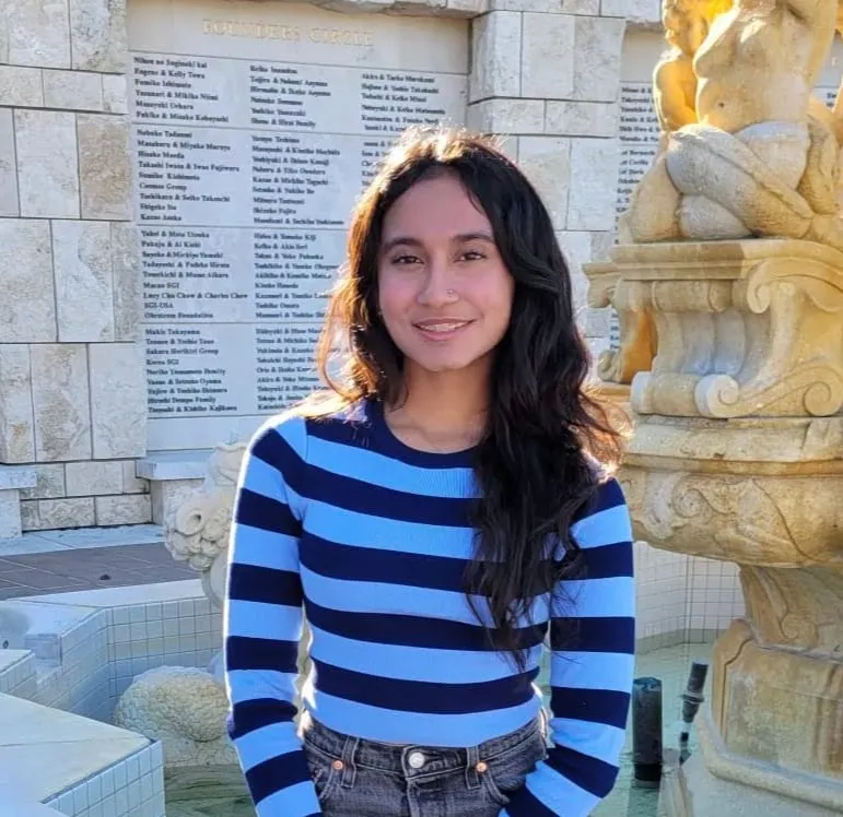 Ira Adnaik in a blue and navy striped sweater and jeans, standing beside a stone fountain and donor wall