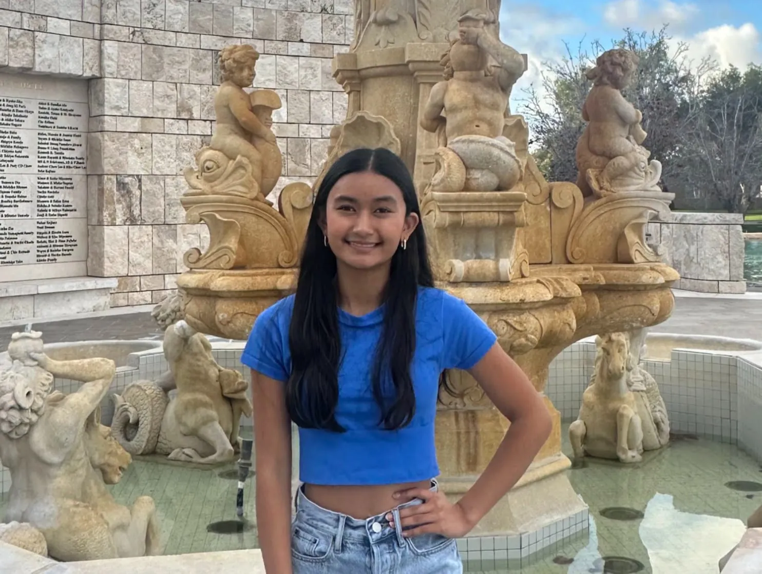 Jayzelle Jahanzeb in front of a tiered stone fountain, wearing a blue cropped T-shirt and light jeans with one hand on her hip