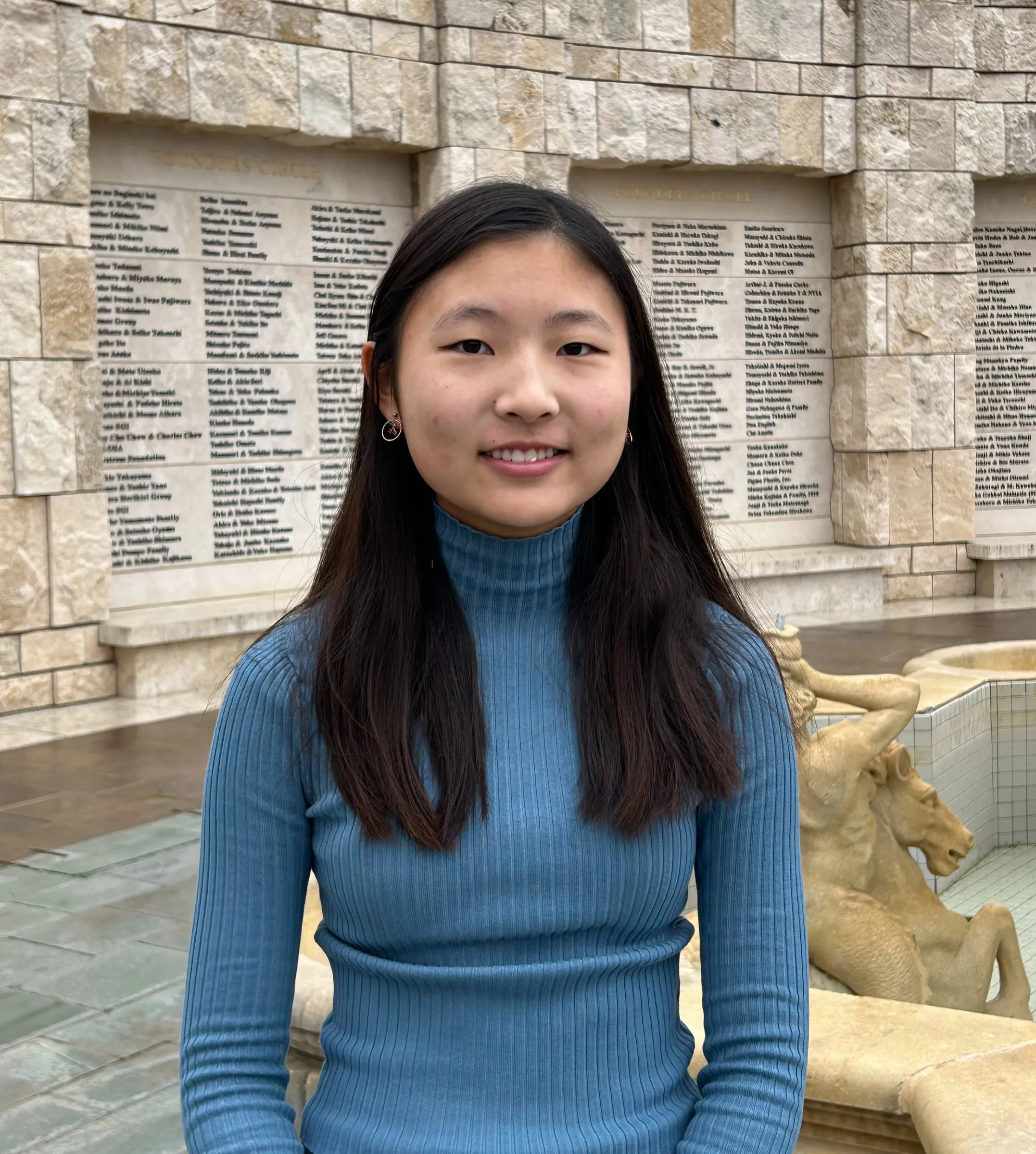 Olive Kim in a blue ribbed turtleneck, standing in front of the Founders Circle donor wall and fountain
