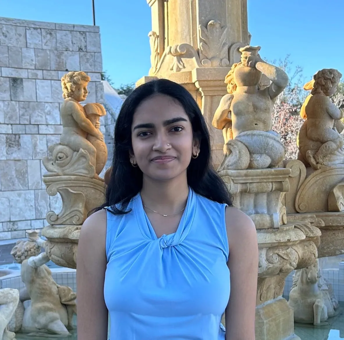 Sanvi Shah in front of an ornate stone fountain with cherub statues, wearing a light blue sleeveless top