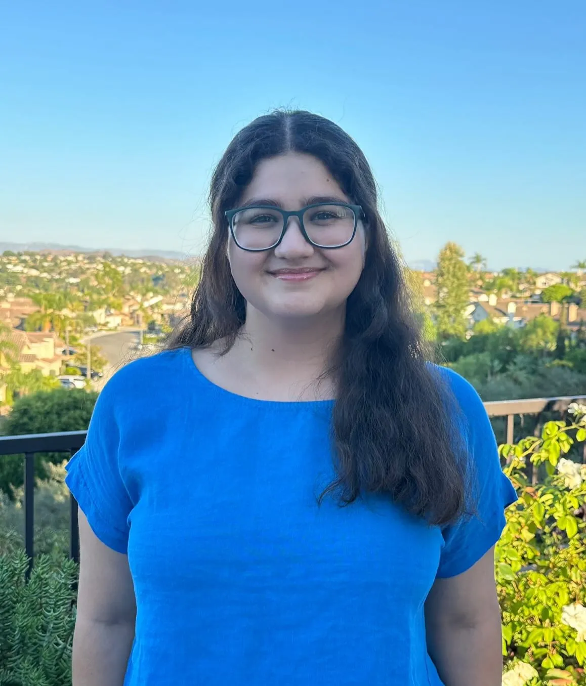 Shiley Moeinzadeh on a balcony overlooking hillside houses, wearing glasses and a bright blue linen top