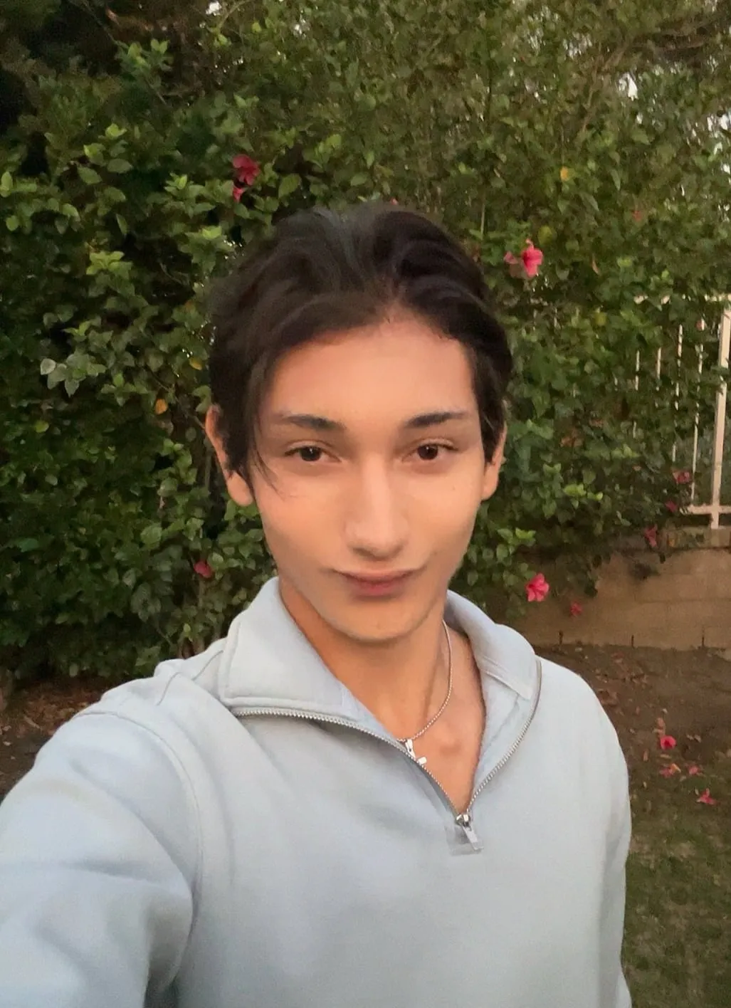 Windy Muccini taking a selfie in front of a flowering bougainvillea hedge, wearing a light blue quarter-zip sweatshirt