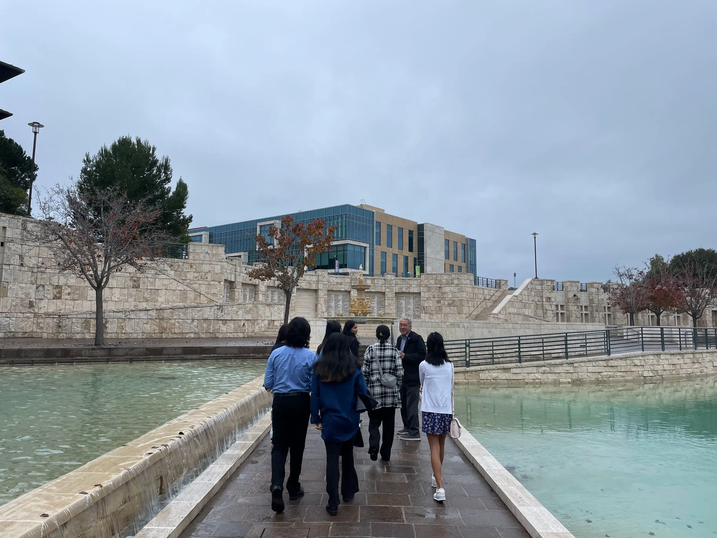 Council members walk single file along a stone causeway between two shallow pools towards their advisor, with the Soka University campus rising behind them